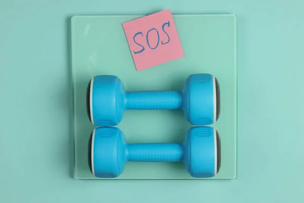 Sport, fitness concept. Losing weight. Floor scales with memo piece of paper with the word SOS and dumbbells on a blue background. Top view.
