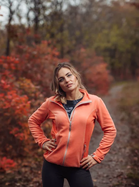 Sonbahar ormanında ağaçlarda kızıl yapraklar olan spor giysili genç bir kadın resmi.