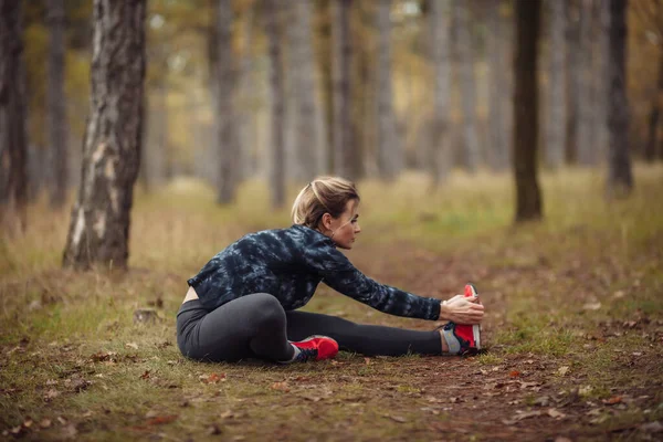 Genç bir kadının sonbahar ormanında otururken koşmadan önce bacak esnemesi.