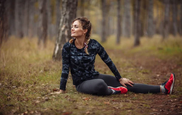 Genç bir kadının sonbahar ormanında otururken koşmadan önce bacak esnemesi.