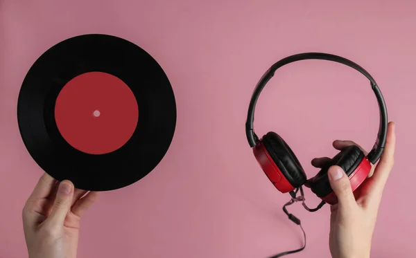 Female hands hold wired headphones with a vinyl record on pink pastel background. Retro style, DJ. Top view, minimalistic music concept.