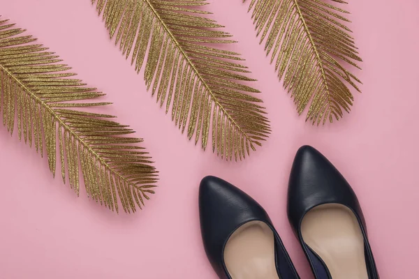 Minimalistic fashion still life. Classic heeled shoes with golden palm leaves on pink pastel background. Studio shot. Top view