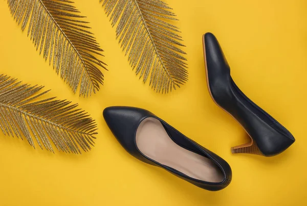 Minimalist moda hala hayattadır. Sarı arka planda altın palmiye yaprakları olan klasik topuklu ayakkabılar. Stüdyo çekimi. Üst görünüm