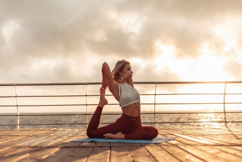 Healthy woman practising yoga on seaside promenade. Sport woman sitting on mat. Eka Pada Rajakapotasana pose