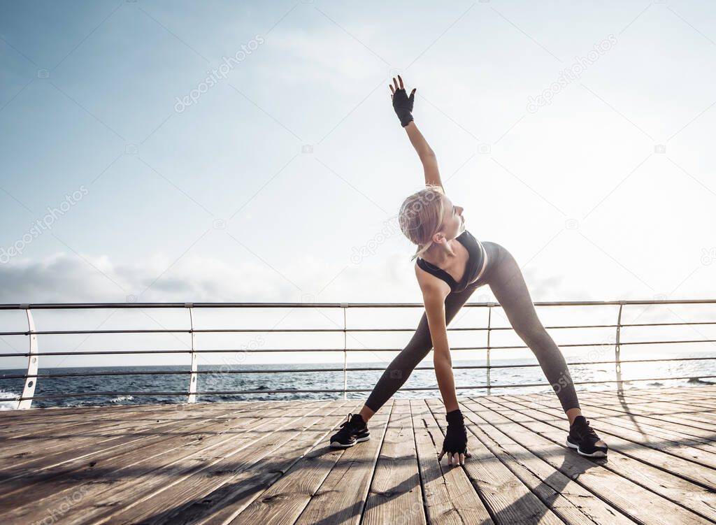 Healthy woman training on seaside promenade. Young attractive woman in sportswear does stretching before exercise on the beach at sunrise. Warm up