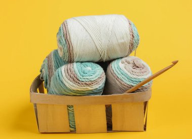 Pastel renklerin yün iplikleri ve sarı arka plandaki sepetin içine tığ işi kanca. Ev Kadınları Hobileri