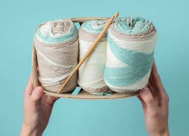 Kadınların elleri pastel renklerin yün ipliklerini tutuyor ve mavi arka planda sepetin içine tığ işi kanca takıyor. Ev Kadınları Hobileri. Üst görünüm