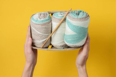 Kadınların elleri pastel renklerin yün ipliklerini tutuyor ve sarı arka planda sepetin içine tığ işi kanca takıyor. Ev Kadınları Hobileri. Üst görünüm