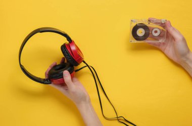 Kadın eli sarı arka planda ses kaseti ve kulaklık tutuyor. Minimalist retro tarzı natürmort hayat. Üst görünüm