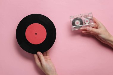 Kadın eli, pembe pastel arka planda ses kaseti ve vinil plak tutuyor. Minimalist retro tarzı natürmort hayat. Üst görünüm
