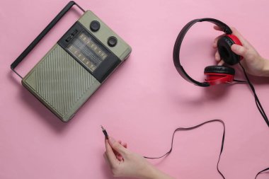 Kadın eli, pembe pastel arka plandaki retro radyo alıcısına tel kulaklık bağlıyor. Üst görünüm