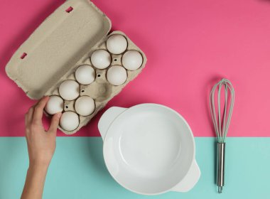 Pişirme süreci. Dişi el, pembe-mavi arka plandaki tepsiden boş bir kase ve çırpıcıyla bir yumurta alır. Üst görünüm