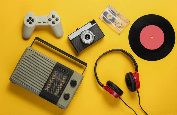 FM radio receiver, retro style wired headphones, vinyl record, gamepad, audio cassette, film camera on yellow background. Top view. 70s