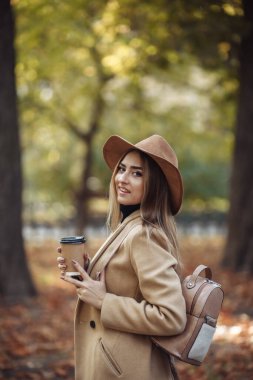 Sonbahar kıyafetleri içinde genç çekici bir kadın ve şehir parkında yürürken kahve içiyor. Şık giyinmiş ve keçeli şapkalı bir kadın. Sonbahar zamanı