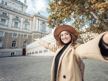 Avrupa kentsel mimarisinin arka planında paltosu ve şapkası olan şık bir turistin selfie portresi. Tatil ve turizm konsepti