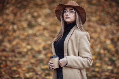 Ceket giyen genç ve çekici bir kadının görüntüsü ve sonbahar parkında düşen yaprakların bulanık arka planında şapkasını hissetti.