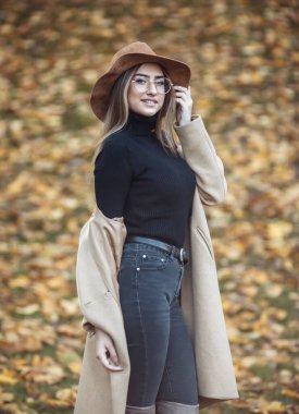 Ceket giyen genç ve çekici bir kadının görüntüsü ve sonbahar parkında düşen yaprakların bulanık arka planında şapkasını hissetti.