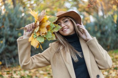 Paltolu ve şapkalı komik genç bir kadın sonbahar parkında bir kucak dolusu yaprak tutuyor. Sonbahar eğlencesi