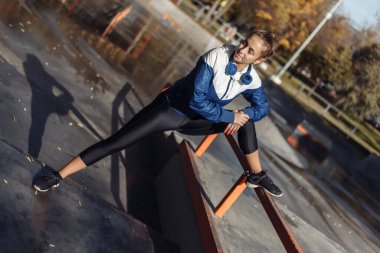 Spor giyim ve kulaklık giyen sıska bir kadın spor parkında antrenman yapmadan önce bacak esneme egzersizi yapıyor.