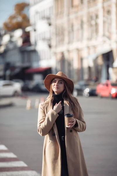 Young stylish woman dressed in a coat and felt hat walks around the city and drinks coffee on the go. Autumn time
