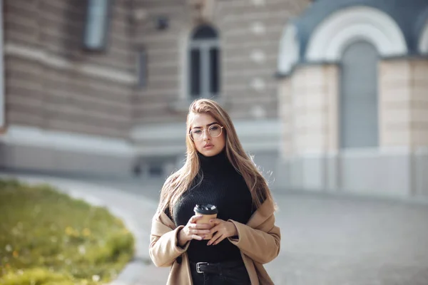穿着时髦外套的年轻女性在城市建筑风格的背景下喝咖啡 — 图库照片