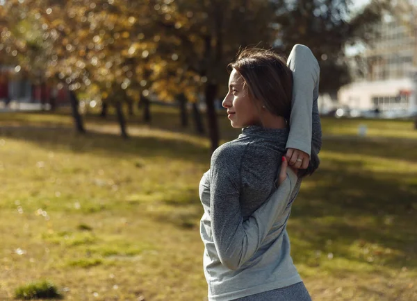 Young sport woman in a sports suit is warming up in the autumn park. Stretching hands before training. Fitness training outdoors