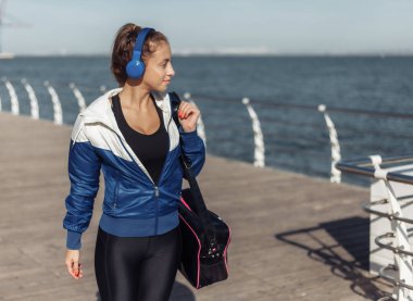 Spor çantalı ve kulaklıklı genç bir kadın güneşli bir günde kumsalda geziniyor.