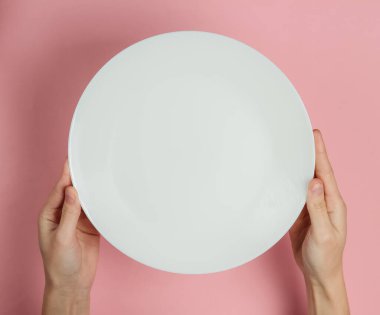 Kadın eli, pembe pastel arka planda boş beyaz bir tabak tutuyor. Üst görünüm.