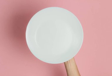 Kadın eli, pembe pastel arka planda boş beyaz bir tabak tutuyor. Üst görünüm.