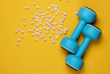 Plastik halterler ve sarı arka planda bir sürü marşmelov. Spor konsepti. Üst görünüm