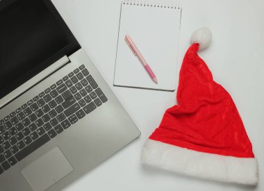 Noel Baba şapkalı dizüstü bilgisayar, beyaz arka planda not defteri. Noel Baba 'ya mektup, istek listesi.
