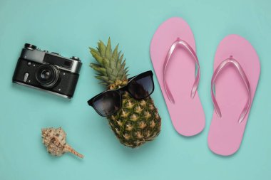 Yaz dönemi. Eğlence ve mizah. Plaj tatili konsepti, seyahat. Güneş gözlüklü ananas, parmak arası terlik, mavi pastel arka planda kamera. Üst görünüm