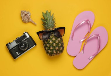 Yaz dönemi. Eğlence ve mizah. Plaj tatili konsepti, seyahat. Güneş gözlüklü ananas, parmak arası terlik, sarı arka planda kamera. Üst görünüm