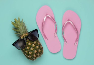 Yaz dönemi. Eğlence ve mizah. Plaj tatili kavramı. Güneş gözlüklü ananas, mavi pastel arka planda parmak arası terlik. Üst görünüm