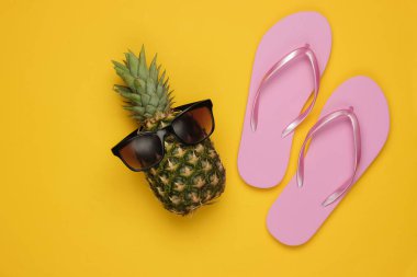 Yaz dönemi. Eğlence ve mizah. Plaj tatili konsepti, seyahat. Güneş gözlüklü ananas, sarı arka planda parmak arası terlik. Üst görünüm