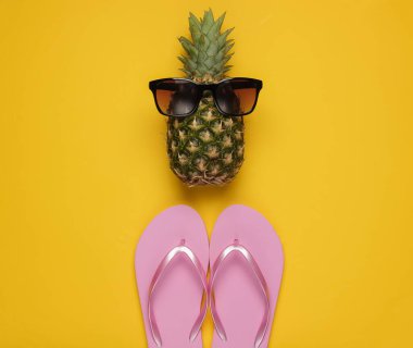 Yaz dönemi. Eğlence ve mizah. Plaj tatili kavramı. Güneş gözlüklü ananas, sarı arka planda parmak arası terlik. Üst görünüm
