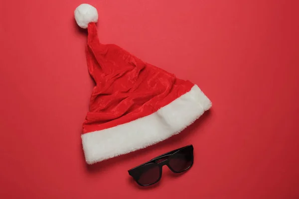 Santa hat and sunglasses on red background. New Year, Christmas holiday in warm sunny places. Beach vacation. Travel concept