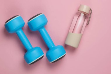 Düz konsept, sağlıklı yaşam tarzı, spor ve zindelik. Dambıllar, pembe pastel arka planda bir şişe su. Üst görünüm