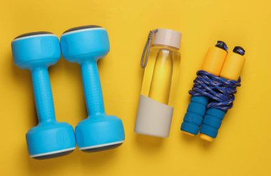 Düz konsept, sağlıklı yaşam tarzı, spor ve zindelik. Dambıllar, bir şişe su, sarı arka planda ip atlama. Üst görünüm