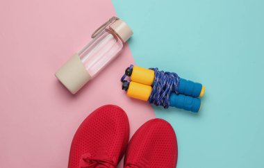 Düz konsept, sağlıklı yaşam tarzı, spor ve zindelik. Koşu için spor ayakkabıları, bir şişe su, pembe mavi pastel arka planda atlama ipi. Üst görünüm