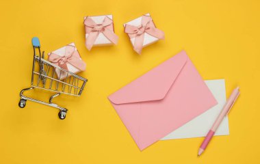 Mektup ve kalemle zarf, hediye kutuları, sarı arka planda alışveriş arabası. Noel, sevgililer günü, doğum günü. Üst görünüm