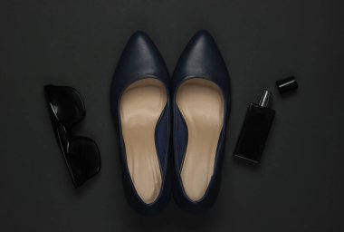 Siyah arka planda kadın aksesuarları. Yüksek topuklu ayakkabılar, parfüm şişesi, güneş gözlüğü. Üst görünüm