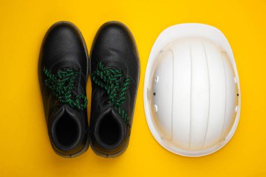 Güvenlik ekipmanları inşaat kaskı, sarı arka planda deri ayakkabılar. Üst görünüm