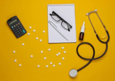 Doktora malzemeleri ve aletler, sarı arka planda hesap makinesi. Sağlık ve tıbbın etkinliğinin ekonomik hesaplanması. Doktor çalışma alanı. Üst görünüm
