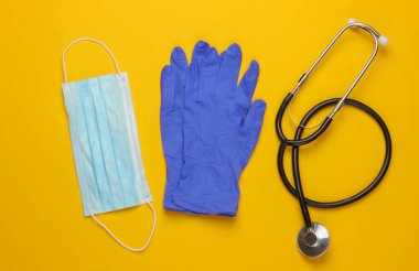 Salgın hastalık. Tıbbi eldiven ve maske, sarı arka planda steteskop. Üst görünüm