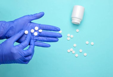 Eldiven içindeki doktor elleri mavi arka planda hapları tutuyor.