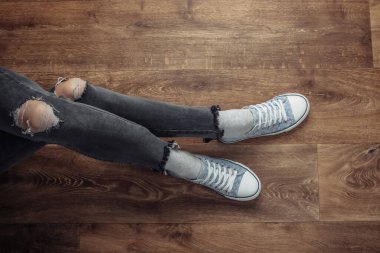 Kadın bacakları yırtık kot pantolon ve tahta zeminde oturan spor ayakkabıları giyer. Üst görünüm