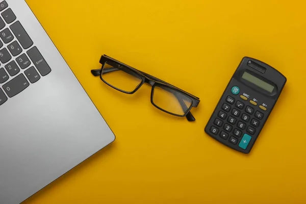 Laptop, glasses, calculator on a yellow background. Top view.