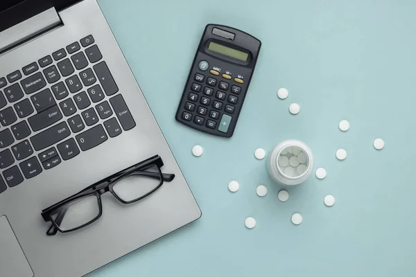 Economic calculation of the cost of treatment, analysis of the cost of medicines. Bottle of pills, laptop, calculator, glasses on blue background. Top view