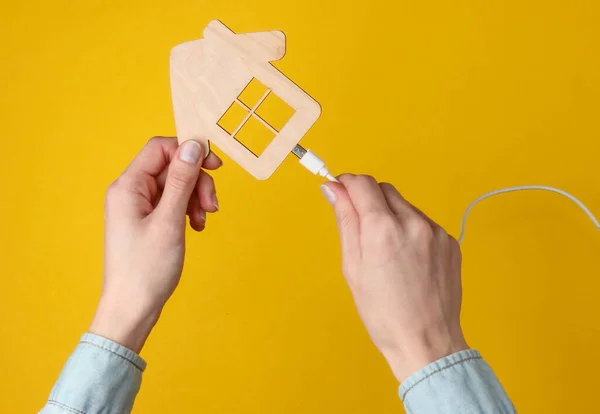 Modern akıllı ev konsepti. Kadın eli, şarj kablosunu sarı arka plandaki evin figürlerine bağlar.